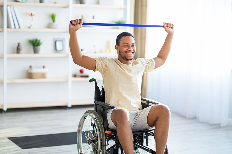 Happy black disabled man in wheelchair doing exercises with rubber band at home. African American handicapped guy working out indoors, keeping in grey shape, enjoying his training