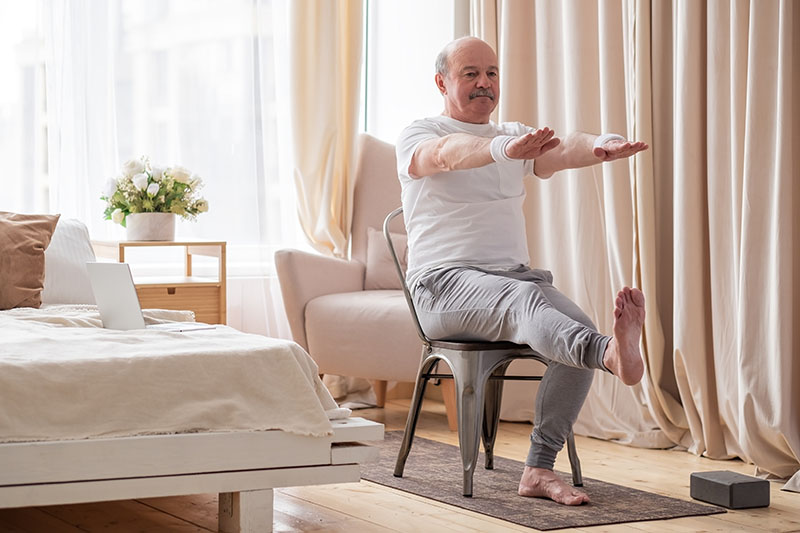 Elderly man practicing yoga asana or sport exercise for legs and hands using chair. Positive mood on sports activities.