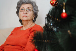 Portrait of lonely sad senior woman with glasses sitting alone at home near xmas tree and looking away. Loneliness in Christmas celebration, self-isolation, lockdown for holiday.